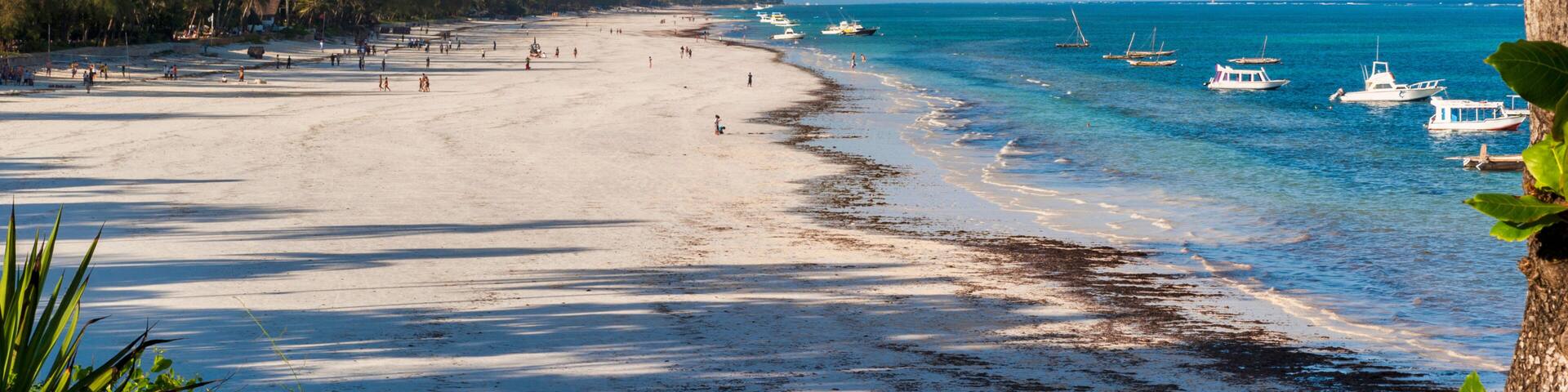 A beach with clear turquoise water, boats, and a sandy shore lined with trees.