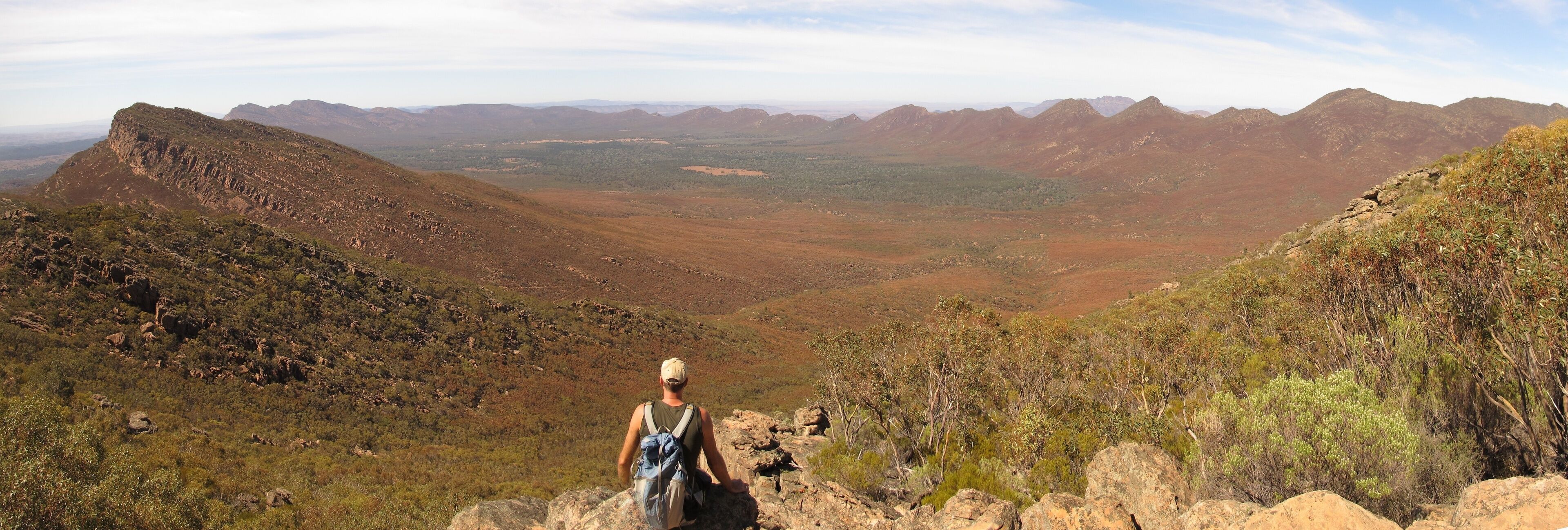 A hiker on a rocky outcrop overlooking a vast mountainous landscape with sparse vegetation.