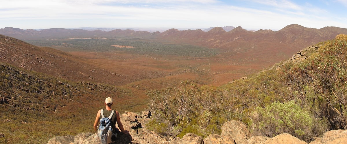 A hiker on a rocky outcrop overlooking a vast mountainous landscape with sparse vegetation.