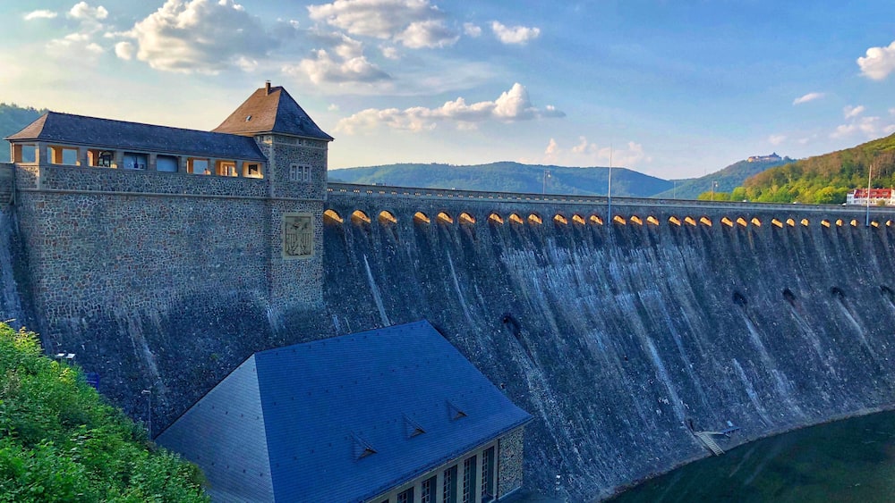Eine historische Steinmauer mit einem Turm, einem Fluss und einer bergigen Landschaft im Hintergrund.