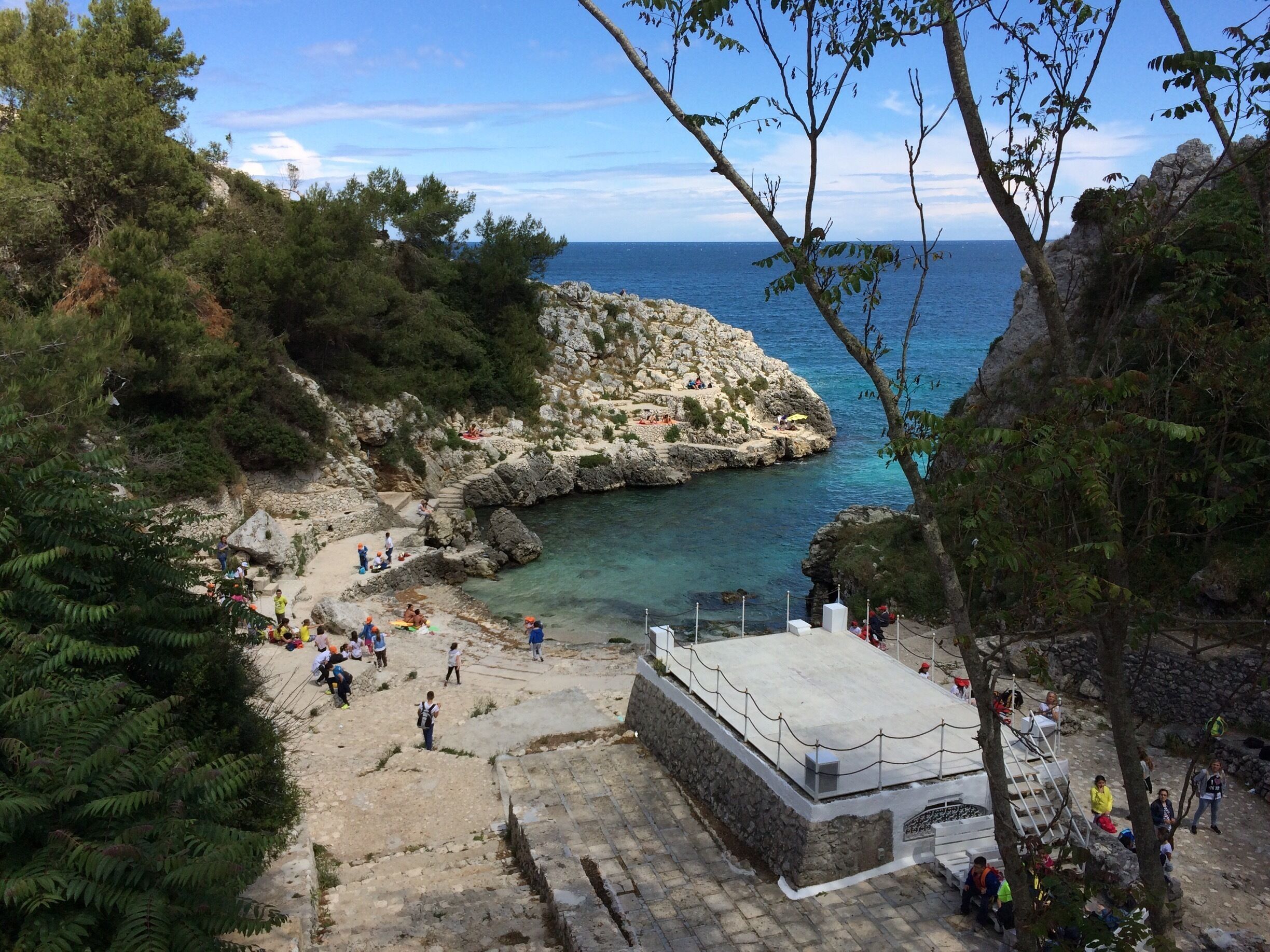 A rocky coastal area with clear blue water, surrounded by dense greenery and a stone structure.
