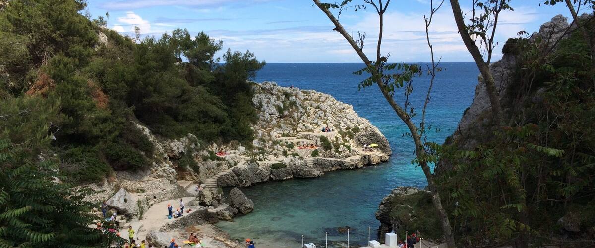 A rocky coastal area with clear blue water, surrounded by dense greenery and a stone structure.