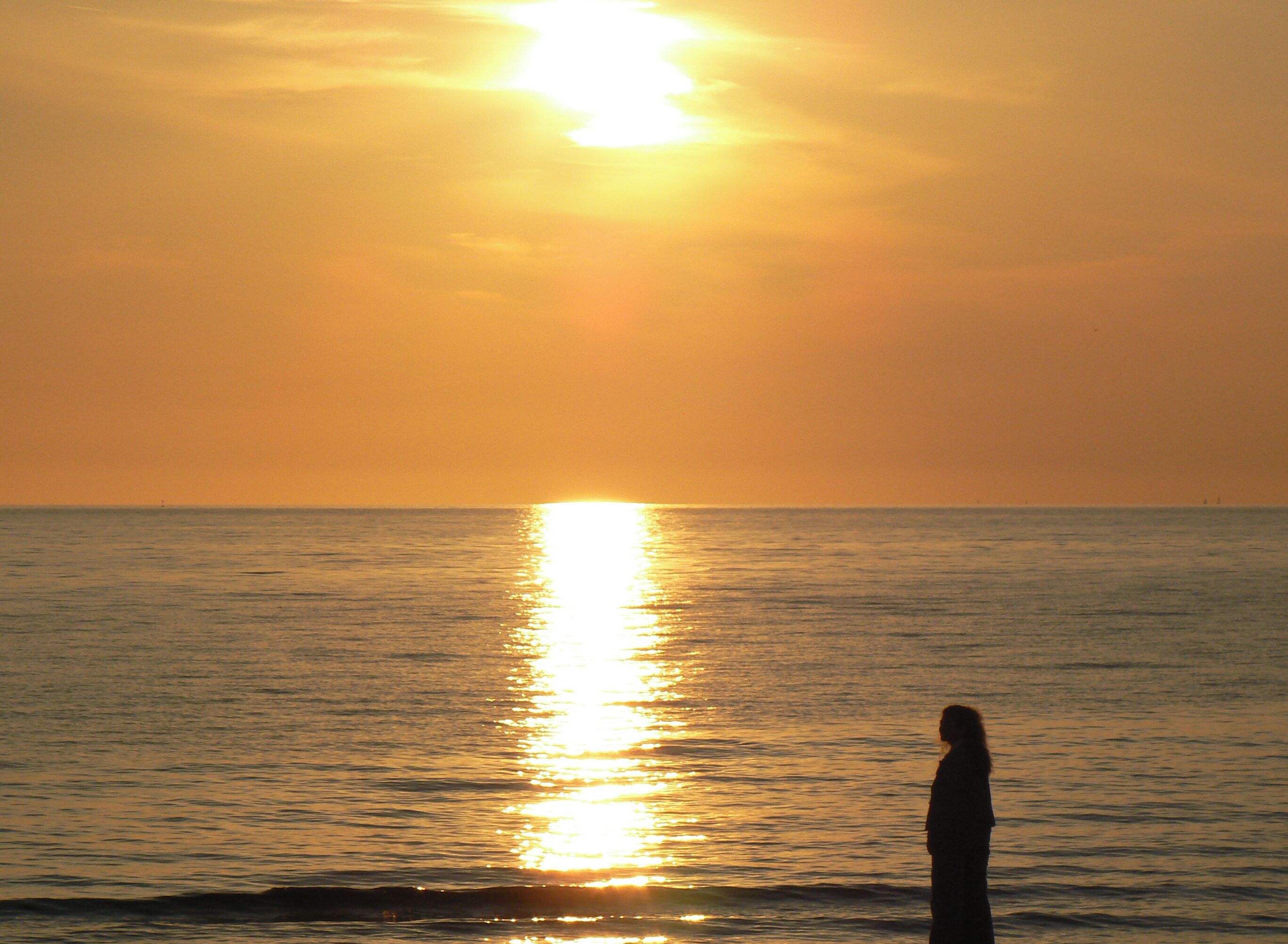 A person standing by the ocean at sunset with the sun reflecting on the water.