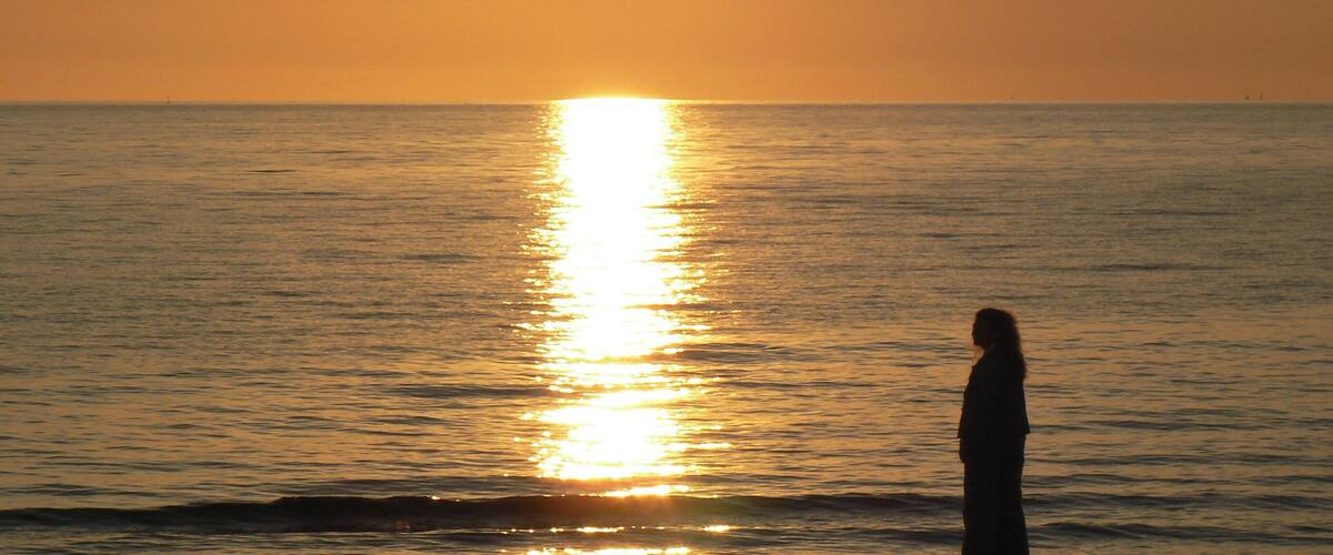 A person standing by the ocean at sunset with the sun reflecting on the water.