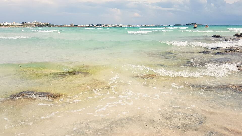 A beach with clear turquoise water, rocky shoreline, and buildings in the distance.