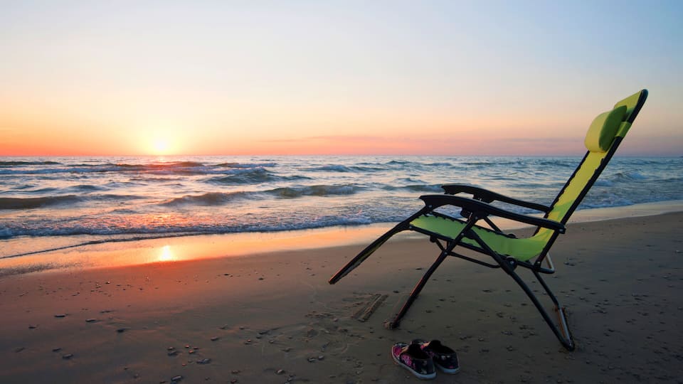 A beach chair and a pair of shoes on the sand near the ocean at sunset.