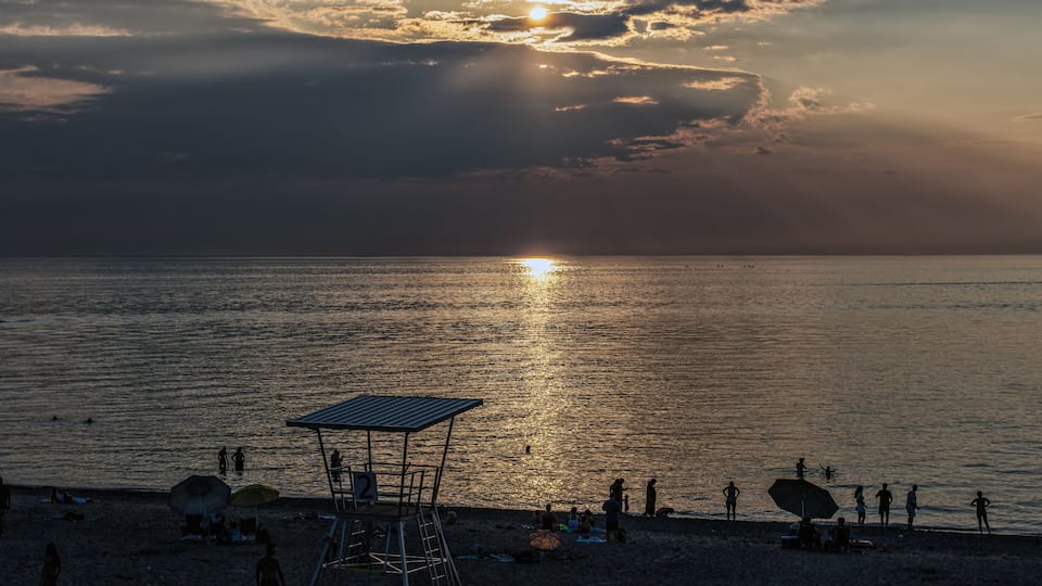 A beach at sunset with the sun reflecting on the water, a lifeguard tower, and people relaxing on the sand.