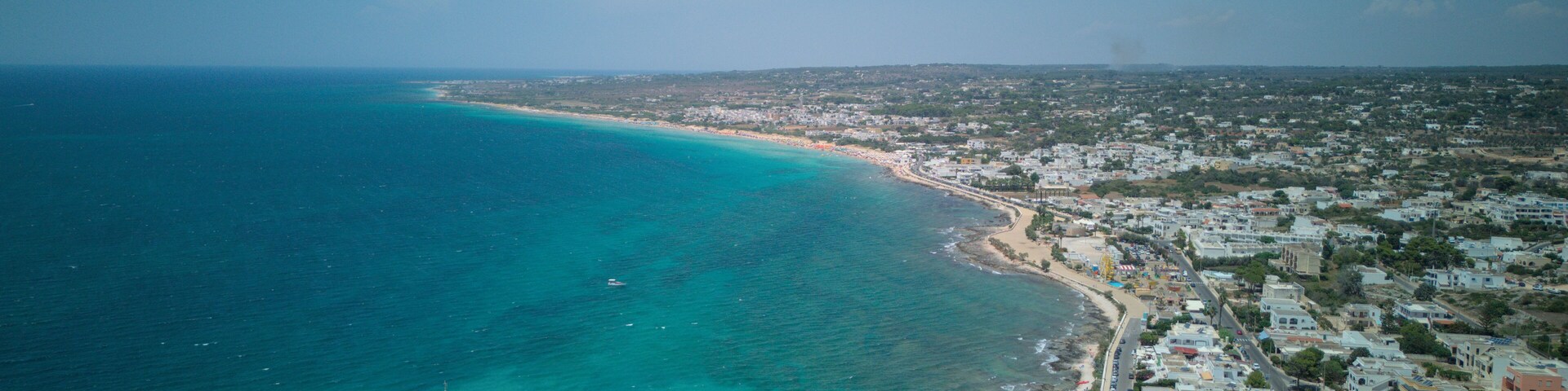 Vista aérea de uma vila costeira com praia, barcos e um molhe rochoso.
