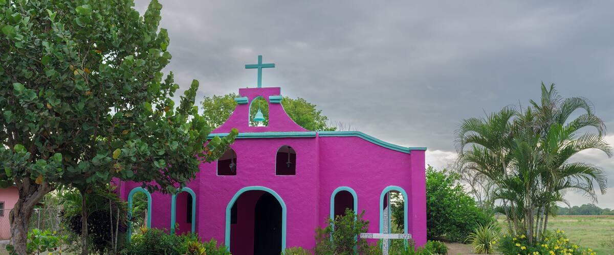 A pink church with a cross on top, surrounded by greenery and palm trees.