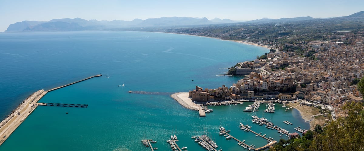 A coastal city with a marina, a fortified castle, and a mountainous backdrop.