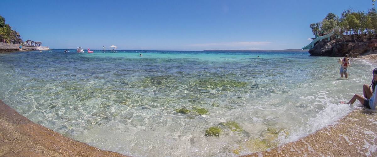 A clear beach with turquoise water, a rocky shoreline, and a few boats in the distance.