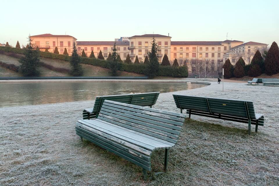 Ein Park mit Bänken an einem Fluss, umgeben von Bäumen und Gebäuden im Hintergrund.