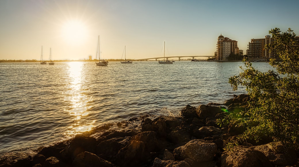 A sunset over a calm river with boats, a bridge, and buildings in the background.