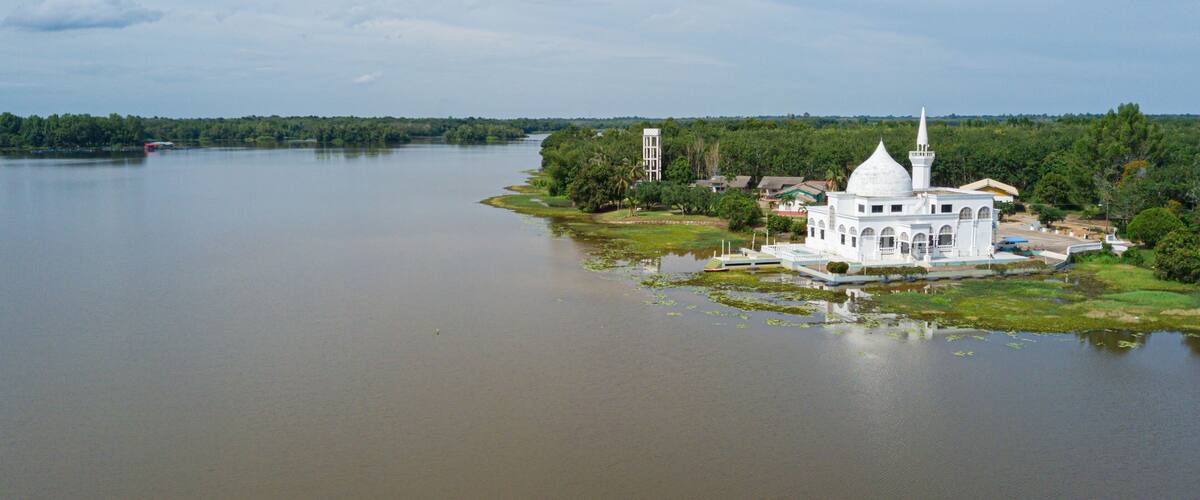 A white mosque with a dome and minaret by a calm river.