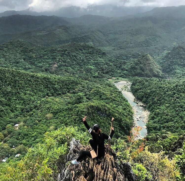 A person standing on a rocky outcrop with arms raised, overlooking a river winding through a dense forested valley.