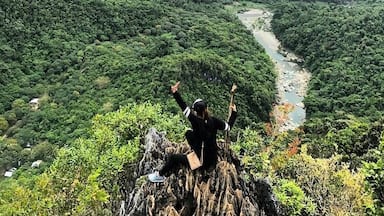 Una persona de pie sobre un afloramiento rocoso, con los brazos levantados, contemplando un río que serpentea a través de un valle cubierto de bosques densos.