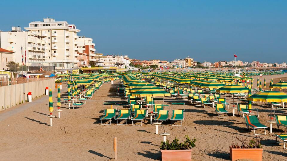 A beach with rows of green and yellow beach chairs and umbrellas, a sandy shore, and buildings in the background.