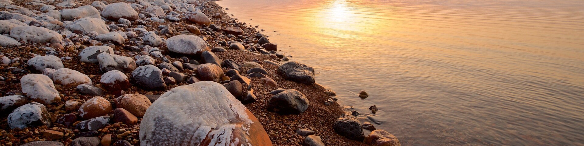 Orilla rocosa con un gran bloque de piedra en primer plano y una masa de agua tranquila que refleja la puesta de sol.