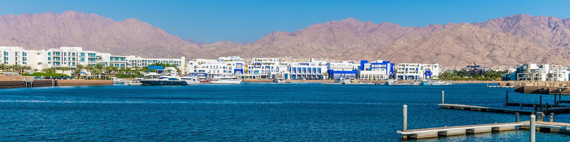 Eine Küstenstadt mit einer Marina, weißen Gebäuden mit blauen Akzenten und Bergen im Hintergrund.