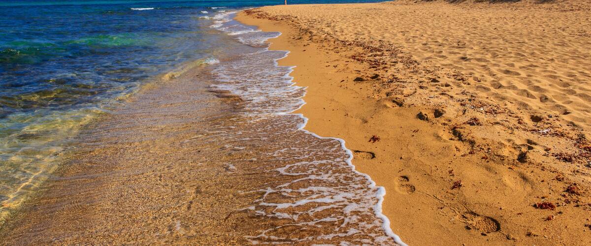 A sandy beach with clear blue water and a person standing in the distance.