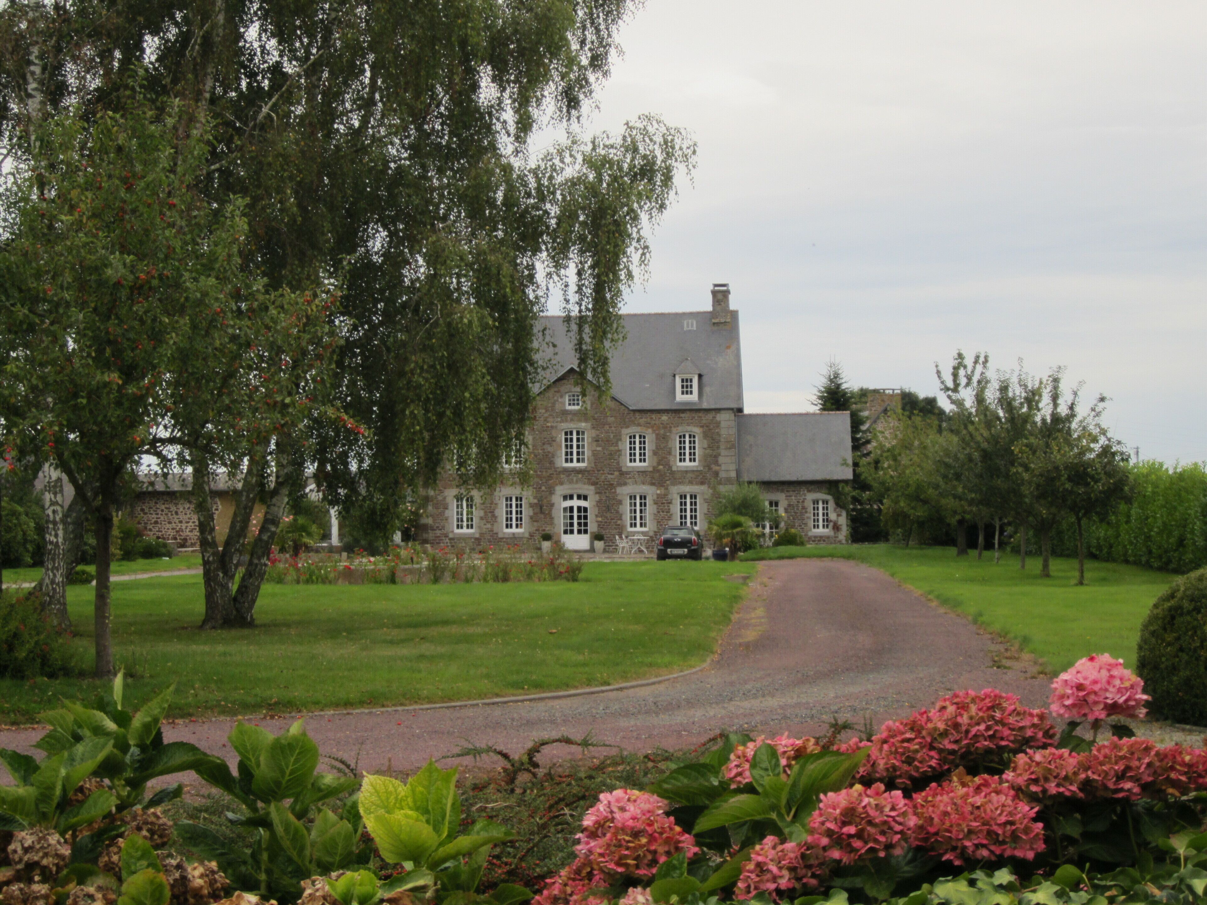 A large stone house with a driveway leading to it, surrounded by lush greenery and flowering plants.