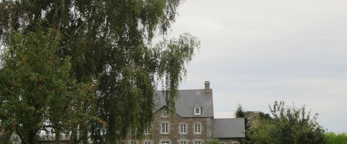 A large stone house with a driveway leading to it, surrounded by lush greenery and flowering plants.