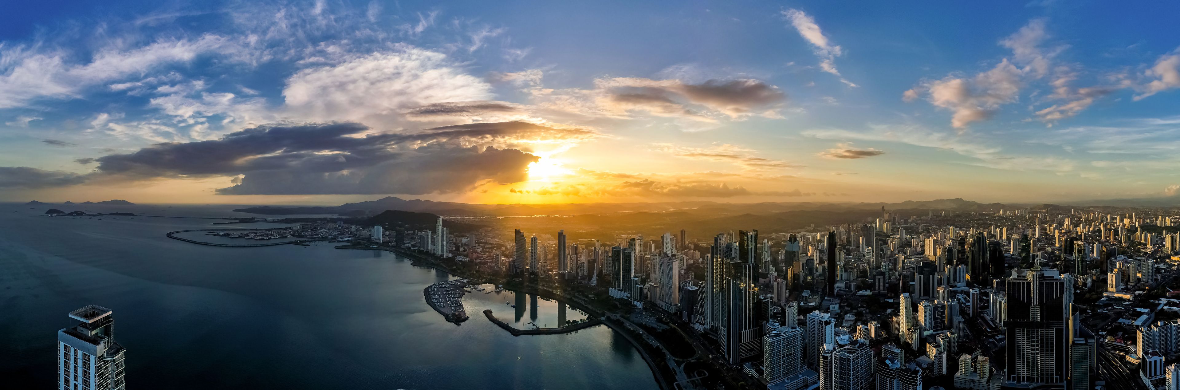 Wide panoramic aerial view of the Panama City skyline and bay silhouetted against a dramatic sunset sky