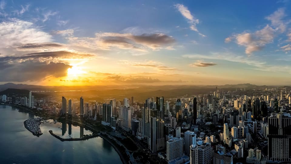 Wide panoramic aerial view of the Panama City skyline and bay silhouetted against a dramatic sunset sky