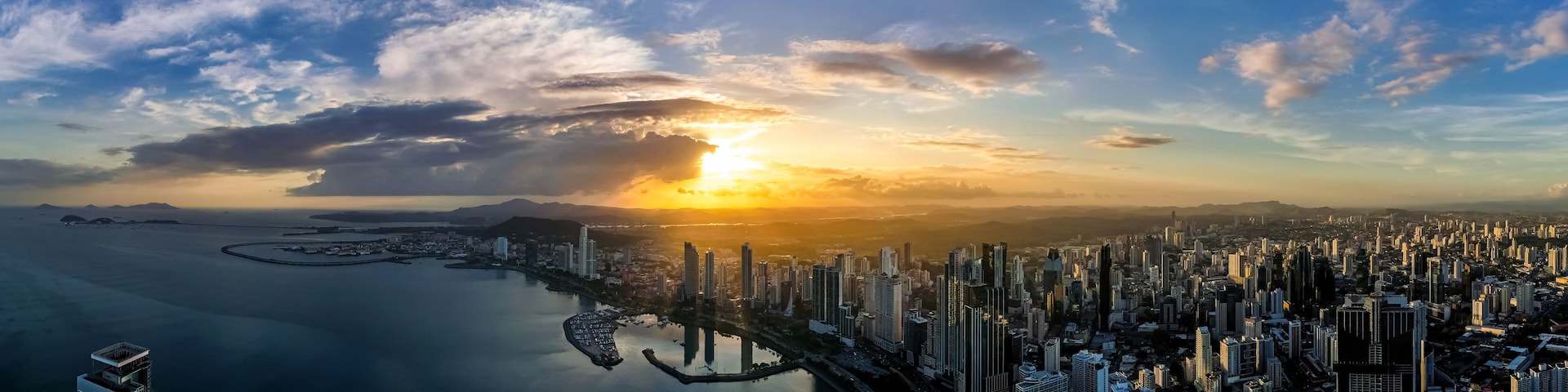 Paisagem costeira com um pôr do sol, edifícios altos e um porto de iates.