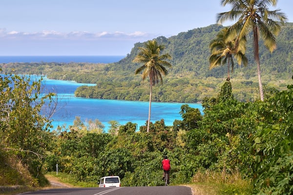 A road with a cyclist and a white van, surrounded by lush greenery and a view of turquoise waters.