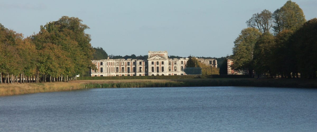 A large, historic building with classical architecture by a calm lake, surrounded by trees.