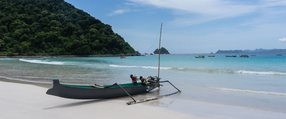 A boat on a beach with clear turquoise water and a forested hill in the background.
