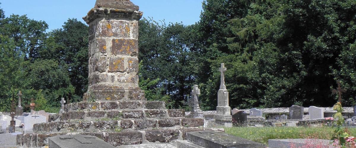 A stone cross atop a weathered monument in a cemetery surrounded by trees.