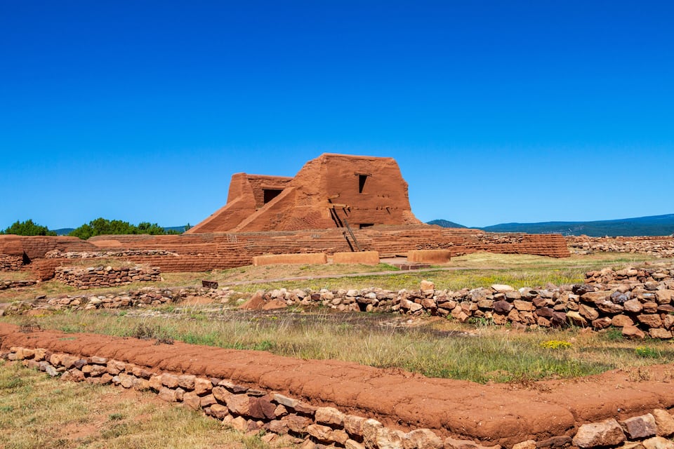 Una estructura antigua de adobe con una amplia sala central y habitaciones más pequeñas adyacentes, rodeada por un muro de piedra y terreno cubierto de hierba.