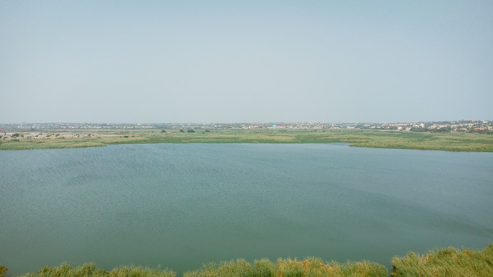 Aerial view of the tranquil lagoon mirroring the vast sky, bordered by lush greenery and distant urban edges, Sakumono, Greater Accra Region, Ghana.