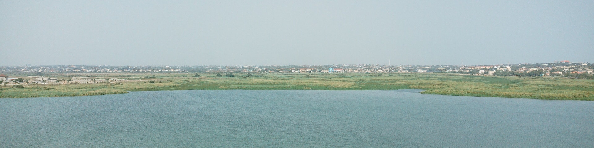 Aerial view of the tranquil lagoon mirroring the vast sky, bordered by lush greenery and distant urban edges, Sakumono, Greater Accra Region, Ghana.