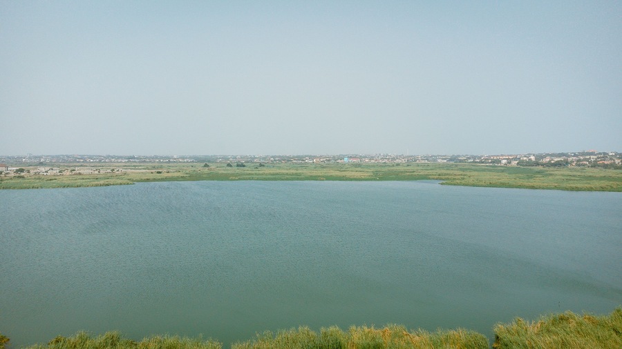 Aerial view of the tranquil lagoon mirroring the vast sky, bordered by lush greenery and distant urban edges, Sakumono, Greater Accra Region, Ghana.