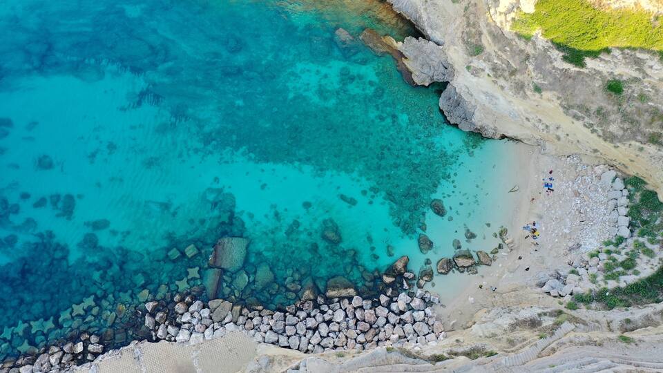 A coastal area with clear turquoise waters, rocky shoreline, and a small group of people on the beach.