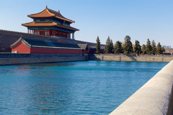 Ein traditionelles chinesisches Gebäude mit rotem Dach und goldenen Verzierungen, gelegen an einem ruhigen Fluss mit einer Steinmauer.