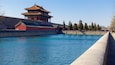 A traditional Chinese building with a red roof and golden accents, situated by a calm river with a stone wall on one side.