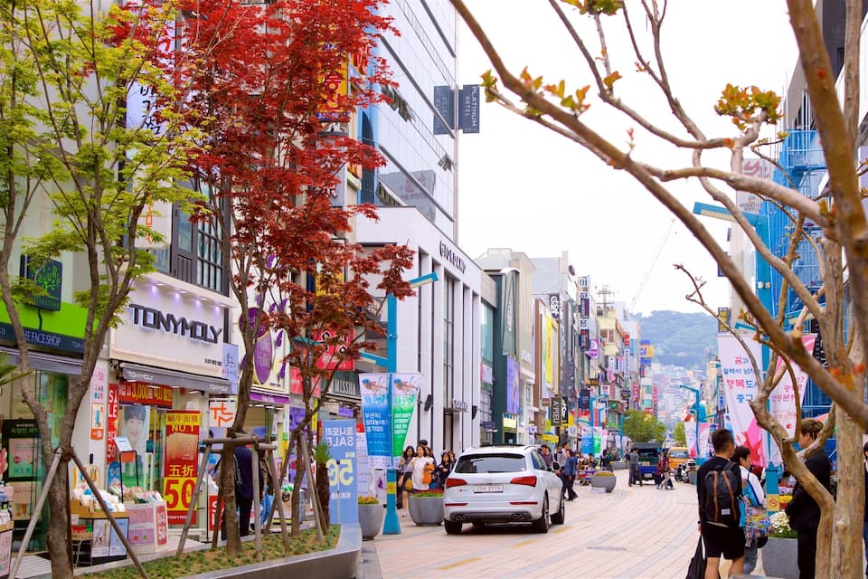 Una concurrida calle urbana bordeada de tiendas y restaurantes, incluyendo una tienda TonyMoly y una tienda Missha de cosméticos.