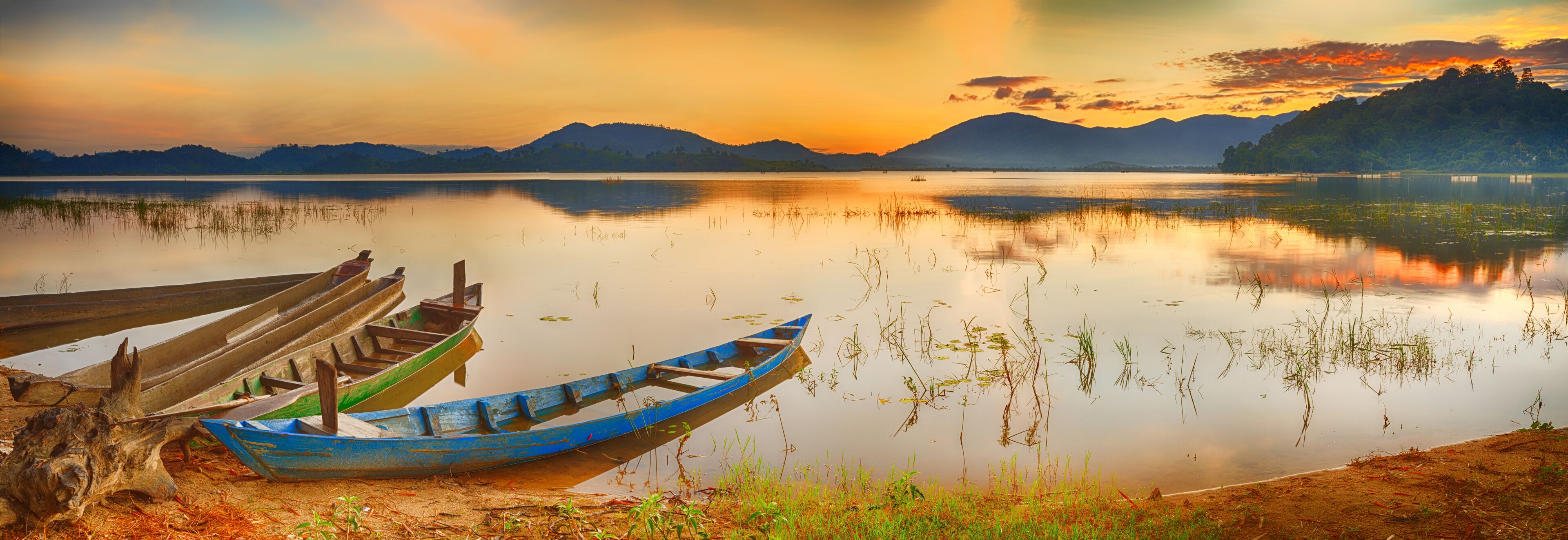 A calm lake at sunset with boats docked on the shore and mountains in the background.