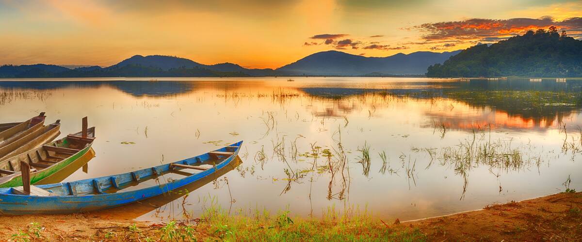 A calm lake at sunset with boats docked on the shore and mountains in the background.