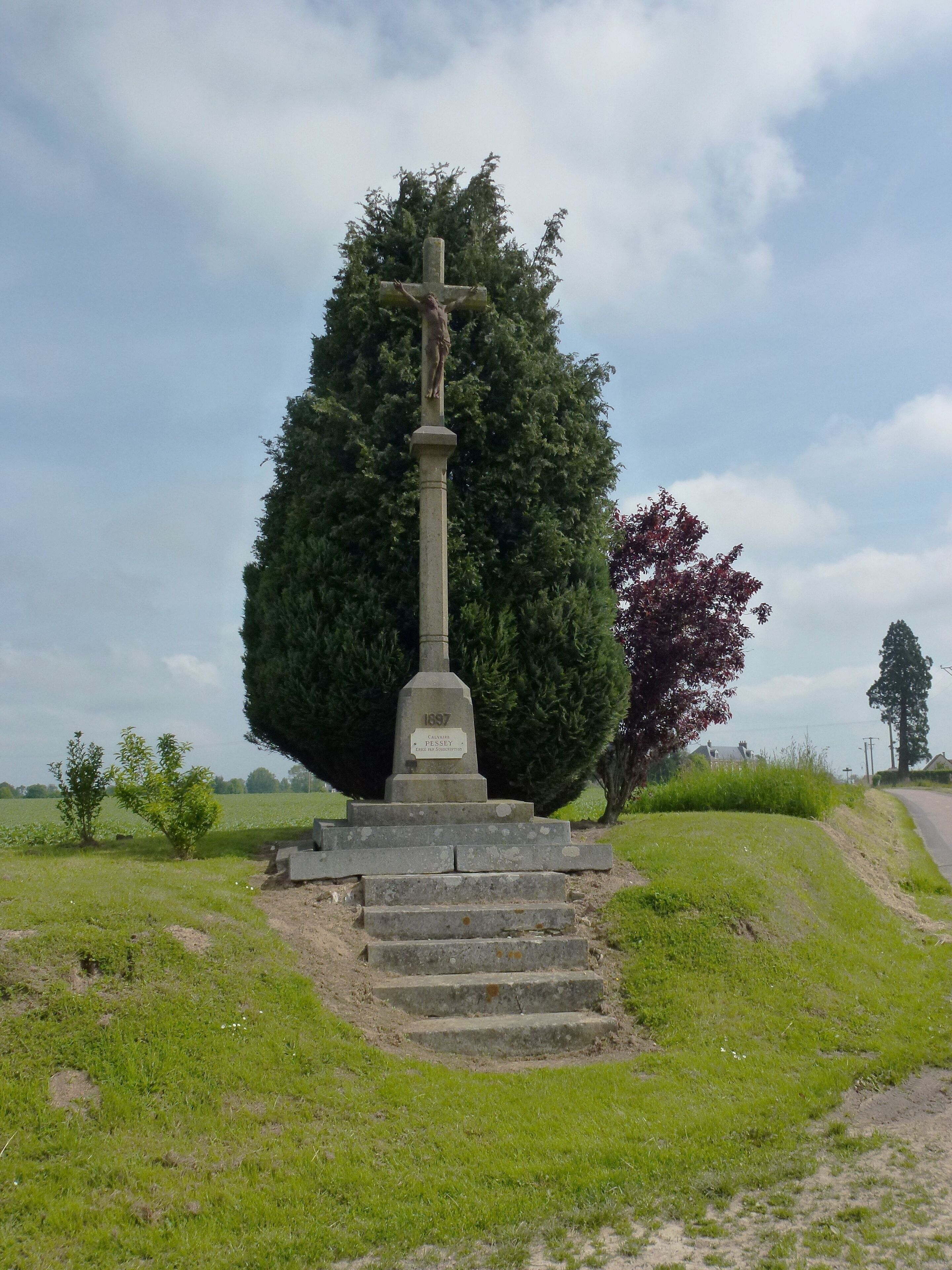 A stone cross monument with inscriptions, surrounded by grass and trees.