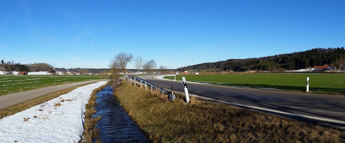 A rural landscape with a road, snow patches, green fields, and a forest in the background.