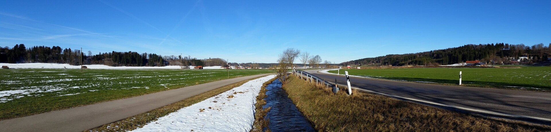 A rural landscape with a road, snow patches, green fields, and a forest in the background.