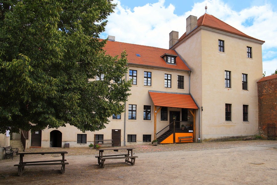 Een historisch gebouw met een rood pannendak, een binnenplaats met banken en een grote boom.