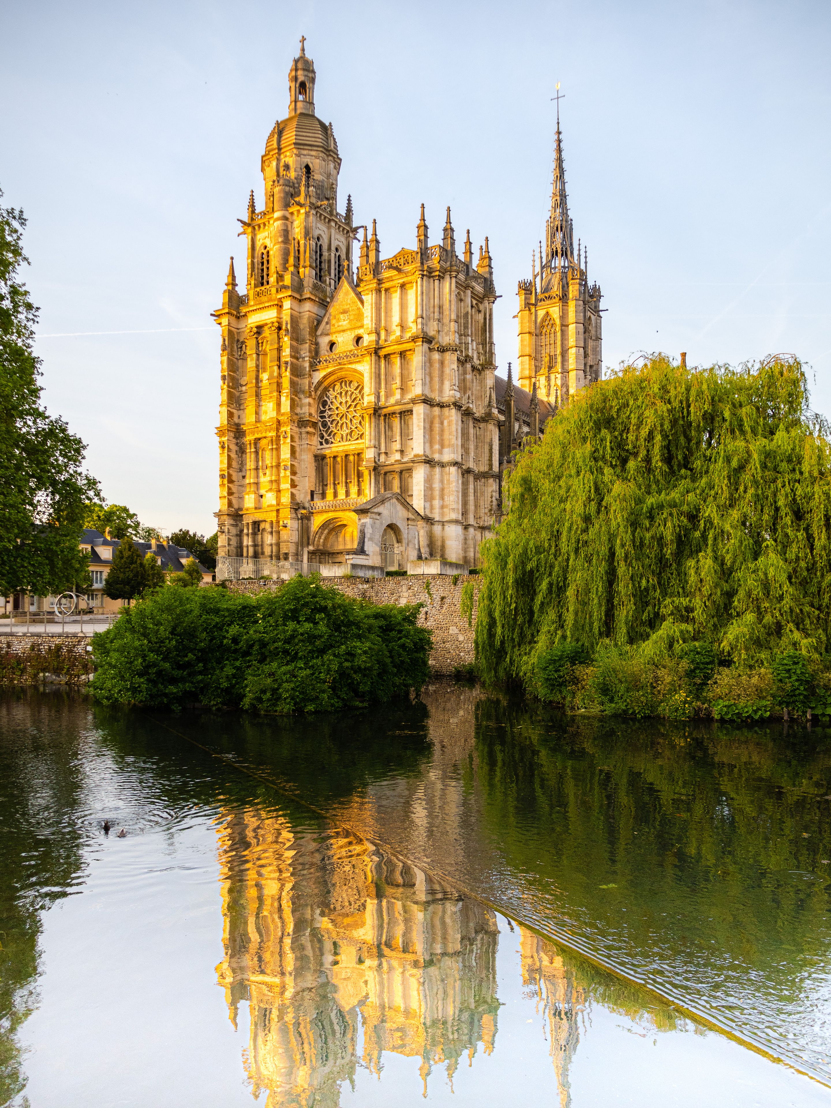 A historic cathedral with intricate architecture, a calm river reflecting its structure, and lush greenery surrounding the area.