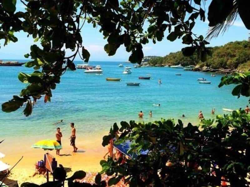 A beach with clear turquoise water, people swimming, and boats in the distance.