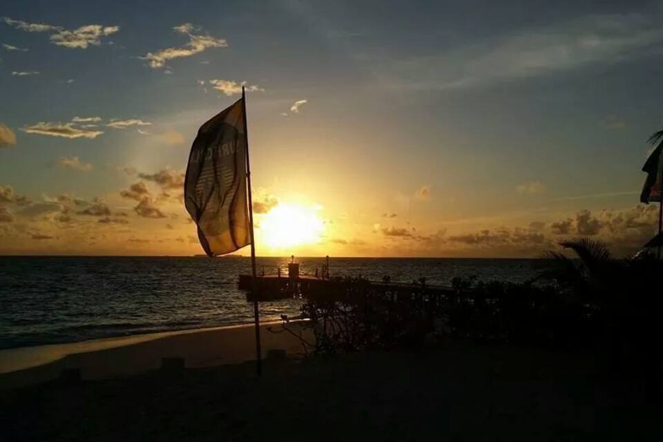 A sunset over a beach with a sailboat and a flag.